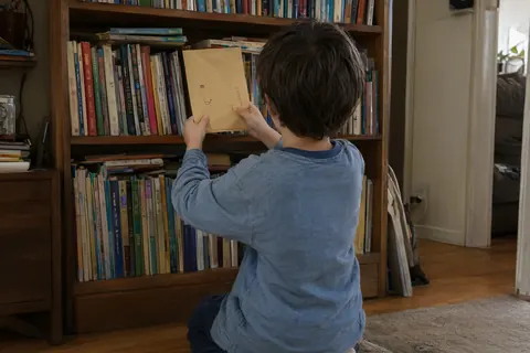 Ein Kind von hinten kniet vor einem Bücherregal und zieht einen Papierumschlag zwischen den Büchern hervor (KI-Bild)